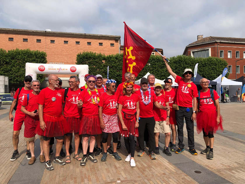 Photo d'un groupe joyeux de participants à la marche nordique. Ils portent tous des t-shirts rouges et certains arborent des tutus rouges ou des colliers de fleurs. Un homme brandit un grand drapeau rouge. Ils posent devant une arche blanche "Nordic Walk" et un bâtiment en briques.