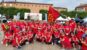 Photo d'un groupe joyeux de participants à la marche nordique. Ils portent tous des t-shirts rouges et certains arborent des tutus rouges ou des colliers de fleurs. Un homme brandit un grand drapeau rouge. Ils posent devant une arche blanche "Nordic Walk" et un bâtiment en briques.