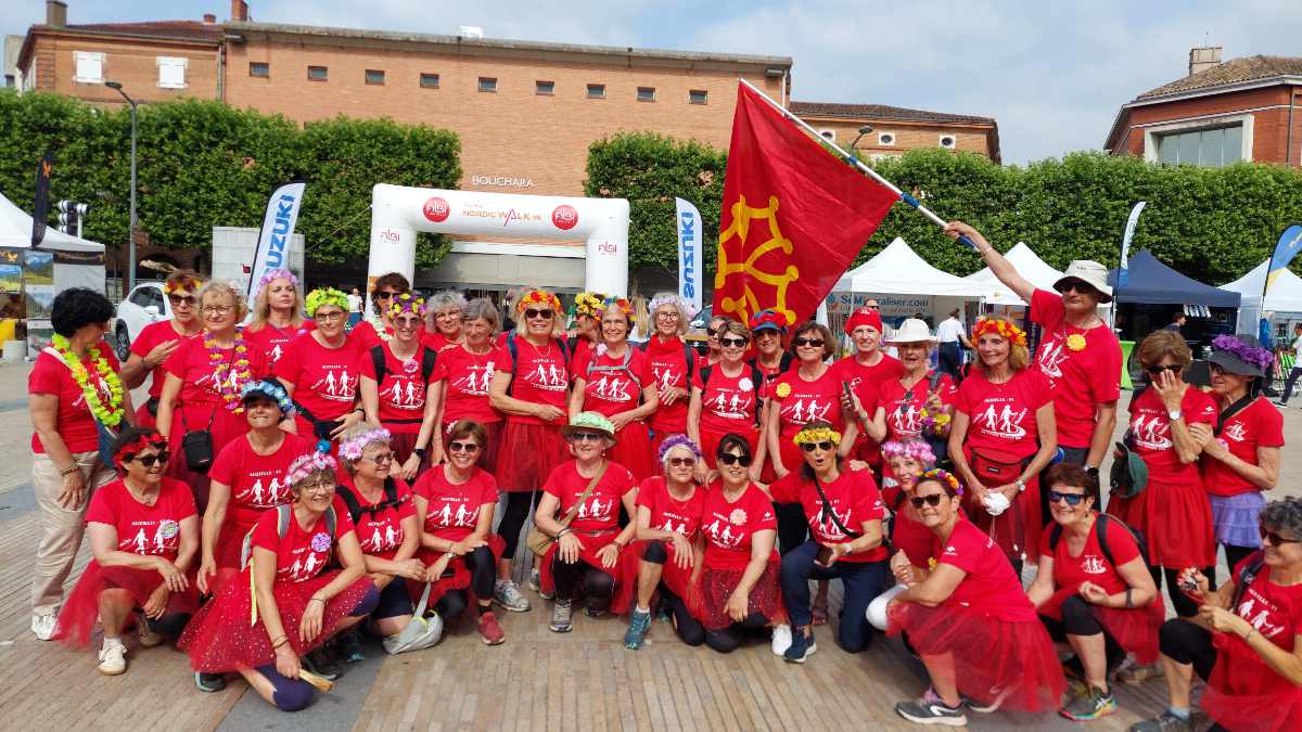 Photo d'un groupe joyeux de participants à la marche nordique. Ils portent tous des t-shirts rouges et certains arborent des tutus rouges ou des colliers de fleurs. Un homme brandit un grand drapeau rouge. Ils posent devant une arche blanche "Nordic Walk" et un bâtiment en briques.
