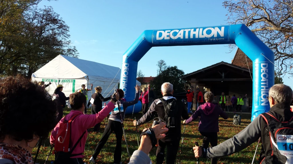 Photo d'un groupe de personnes participant à un événement de marche nordique. Ils se trouvent devant une arche gonflable bleue marquée "DECATHLON". Plusieurs participants sont équipés de bâtons de marche. À l'arrière-plan, on aperçoit une tente blanche et un bâtiment en bois.