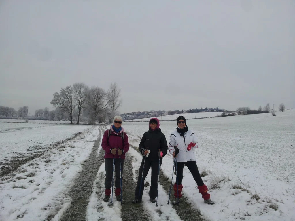 Trois femmes souriantes en tenue de marche hivernale, équipées de bâtons, posent sur un sentier enneigé. Le paysage autour est recouvert de neige, avec des arbres givrés à l'arrière-plan et un village au loin sous un ciel gris.